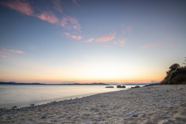 Kumlu bir sahildeki kayaların arasından gün batımına bak. Manzara, Ouranoupoli, Selanik, Orta Makedonya ve Yunanistan kıyılarındaki engin deniz manzarası ile çekildi