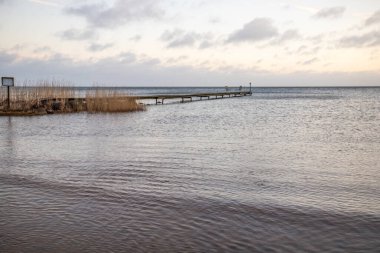 Sunrise over a lake in autumn. A wooden jetty protrudes into the water while grass has established itself on the sandy beach. The lake and beach, located on Lake Vnern in Ns Sannar, Sweden, Scandina clipart