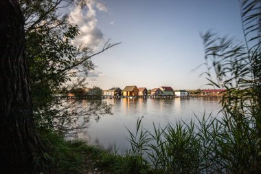 Sudaki ahşap evler. Gün batımında gölde tatil beldesi. Uzun ahşap yollar bireysel evlere çıkıyor. Macaristan, Balaton Gölü, Bokodi 'nin yüzen köyünün manzara fotoğrafı.