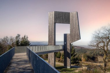 Wooden sculpture with a steel walkway and viewpoint. Landscape shot on a mountain, blue sky fog in the valley. Morning at the Erbeskopf wind sound sculpture, Hilscheid, Rhineland-Palatinate, Germany clipart