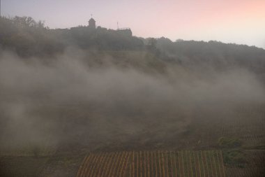 Sonbaharda manzara çekimi. Sisin içinde nehir ve sonbahar bağları arasında bir kasaba dolaşıyor. Tepede bir kale duruyor. Zell Mosel, Hunsrck, Rhineland Palatinate