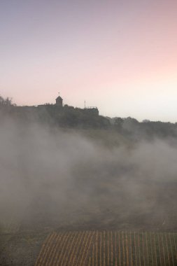 Sonbaharda manzara çekimi. Sisin içinde nehir ve sonbahar bağları arasında bir kasaba dolaşıyor. Tepede bir kale duruyor. Zell Mosel, Hunsrck, Rhineland Palatinate