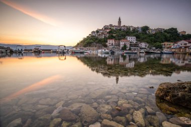 Liman ve sahil, tepenin üstünde eski bir köy. Gemiler suya yansıyor, güneş Adriyatik 'in ufuklarından doğuyor. Vrbnik, Krk, Hırvatistan 'dan güzel manzara görüntüleri