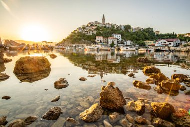 Harbor and beach with an old village on a hill. Boats are reflected in the water, the sun rises on the horizon of the Adriatic. Beautiful landscape shot from Vrbnik, Krk, Croatia clipart