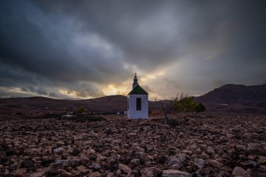 Verimsiz volkanik bir arazide küçük, beyaz ahşap bir şapel. Günbatımı, İspanya 'nın Kanarya Adaları' ndaki Fuerteventura Adası 'ndaki Ermita Protestante de Violante kilisesinin manzara görüntüsü.