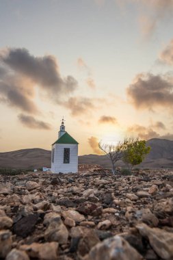 Verimsiz volkanik bir arazide küçük, beyaz ahşap bir şapel. Günbatımı, İspanya 'nın Kanarya Adaları' ndaki Fuerteventura Adası 'ndaki Ermita Protestante de Violante kilisesinin manzara görüntüsü.