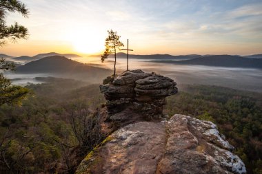 Zirve haçı ve orman manzaralı bir kayanın üzerinde gün doğumu. Doğanın ortasında sisli bir sabah havası. Almanya 'nın Derebeyi Ormanı' ndaki manzara