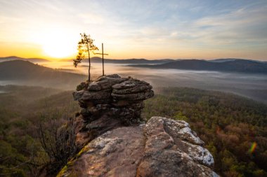Zirve haçı ve orman manzaralı bir kayanın üzerinde gün doğumu. Doğanın ortasında sisli bir sabah havası. Almanya 'nın Derebeyi Ormanı' ndaki manzara
