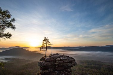 Zirve haçı ve orman manzaralı bir kayanın üzerinde gün doğumu. Doğanın ortasında sisli bir sabah havası. Almanya 'nın Derebeyi Ormanı' ndaki manzara