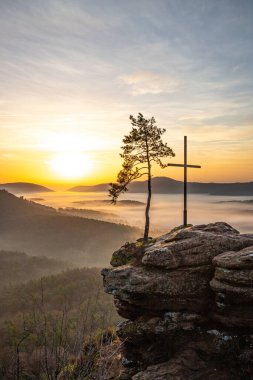 Zirve haçı ve orman manzaralı bir kayanın üzerinde gün doğumu. Doğanın ortasında sisli bir sabah havası. Almanya 'nın Derebeyi Ormanı' ndaki manzara