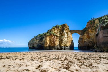 Kumsaldaki sarı kayalar ve uçurumlar. Eski Pinhao Fort Köprüsü 'nün iki uçurumu birleştirdiği deniz kenarında bir koy. Praia da Caldeira plajı, Lagos. Algarve, Portekiz