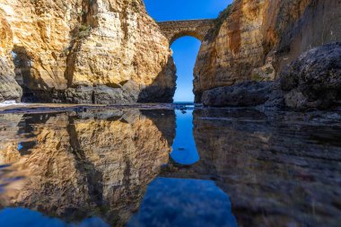 Kumsaldaki sarı kayalar ve uçurumlar. Eski Pinhao Fort Köprüsü 'nün iki uçurumu birleştirdiği deniz kenarında bir koy. Praia da Caldeira plajı, Lagos. Algarve, Portekiz