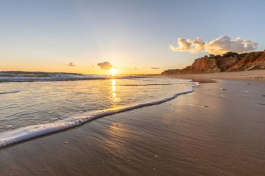 Kızıl kayalıkları ve kayalıkları olan güzel kumlu bir sahil. Günbatımında Praia do Poo Velho, Praia da Falsia, Algarve, Portekiz 'de Atlantik kıyısında sığ dalgalarla birlikte