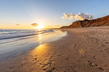 Kızıl kayalıkları ve kayalıkları olan güzel kumlu bir sahil. Günbatımında Praia do Poo Velho, Praia da Falsia, Algarve, Portekiz 'de Atlantik kıyısında sığ dalgalarla birlikte