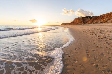 Kızıl kayalıkları ve kayalıkları olan güzel kumlu bir sahil. Günbatımında Praia do Poo Velho, Praia da Falsia, Algarve, Portekiz 'de Atlantik kıyısında sığ dalgalarla birlikte