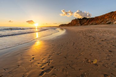 Kızıl kayalıkları ve kayalıkları olan güzel kumlu bir sahil. Günbatımında Praia do Poo Velho, Praia da Falsia, Algarve, Portekiz 'de Atlantik kıyısında sığ dalgalarla birlikte