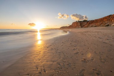 Kızıl kayalıkları ve kayalıkları olan güzel kumlu bir sahil. Günbatımında Praia do Poo Velho, Praia da Falsia, Algarve, Portekiz 'de Atlantik kıyısında sığ dalgalarla birlikte