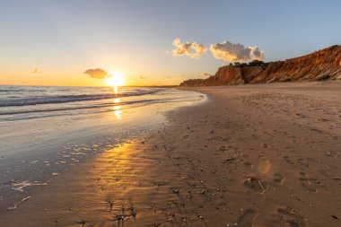 Kızıl kayalıkları ve kayalıkları olan güzel kumlu bir sahil. Günbatımında Praia do Poo Velho, Praia da Falsia, Algarve, Portekiz 'de Atlantik kıyısında sığ dalgalarla birlikte