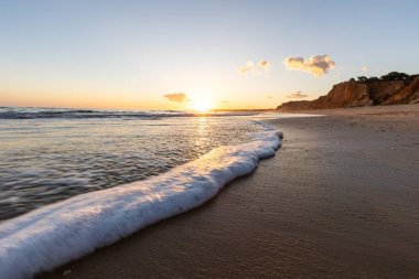 Kızıl kayalıkları ve kayalıkları olan güzel kumlu bir sahil. Günbatımında Praia do Poo Velho, Praia da Falsia, Algarve, Portekiz 'de Atlantik kıyısında sığ dalgalarla birlikte
