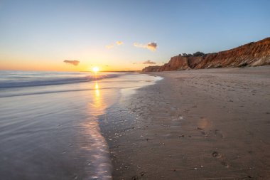 Kızıl kayalıkları ve kayalıkları olan güzel kumlu bir sahil. Günbatımında Praia do Poo Velho, Praia da Falsia, Algarve, Portekiz 'de Atlantik kıyısında sığ dalgalarla birlikte