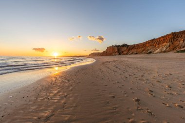 Kızıl kayalıkları ve kayalıkları olan güzel kumlu bir sahil. Günbatımında Praia do Poo Velho, Praia da Falsia, Algarve, Portekiz 'de Atlantik kıyısında sığ dalgalarla birlikte
