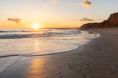 Kızıl kayalıkları ve kayalıkları olan güzel kumlu bir sahil. Günbatımında Praia do Poo Velho, Praia da Falsia, Algarve, Portekiz 'de Atlantik kıyısında sığ dalgalarla birlikte