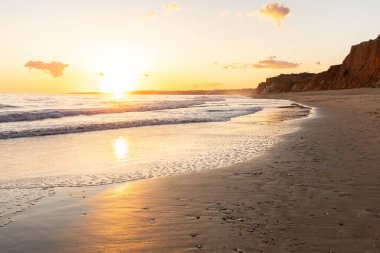 Kızıl kayalıkları ve kayalıkları olan güzel kumlu bir sahil. Günbatımında Praia do Poo Velho, Praia da Falsia, Algarve, Portekiz 'de Atlantik kıyısında sığ dalgalarla birlikte