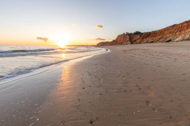 Kızıl kayalıkları ve kayalıkları olan güzel kumlu bir sahil. Günbatımında Praia do Poo Velho, Praia da Falsia, Algarve, Portekiz 'de Atlantik kıyısında sığ dalgalarla birlikte