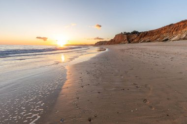 Kızıl kayalıkları ve kayalıkları olan güzel kumlu bir sahil. Günbatımında Praia do Poo Velho, Praia da Falsia, Algarve, Portekiz 'de Atlantik kıyısında sığ dalgalarla birlikte