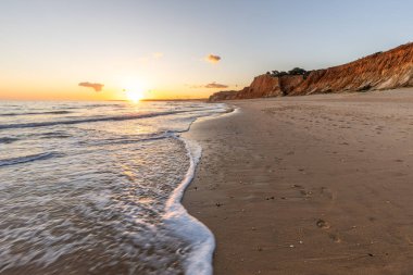 Kızıl kayalıkları ve kayalıkları olan güzel kumlu bir sahil. Günbatımında Praia do Poo Velho, Praia da Falsia, Algarve, Portekiz 'de Atlantik kıyısında sığ dalgalarla birlikte