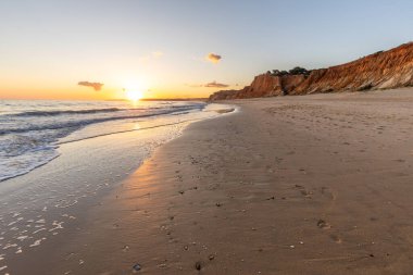 Kızıl kayalıkları ve kayalıkları olan güzel kumlu bir sahil. Günbatımında Praia do Poo Velho, Praia da Falsia, Algarve, Portekiz 'de Atlantik kıyısında sığ dalgalarla birlikte