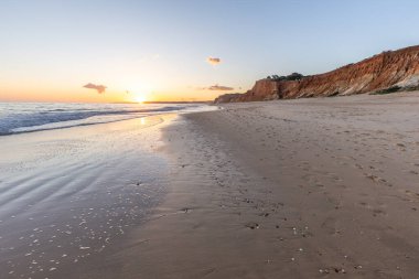 Kızıl kayalıkları ve kayalıkları olan güzel kumlu bir sahil. Günbatımında Praia do Poo Velho, Praia da Falsia, Algarve, Portekiz 'de Atlantik kıyısında sığ dalgalarla birlikte