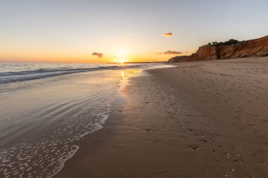 Kızıl kayalıkları ve kayalıkları olan güzel kumlu bir sahil. Günbatımında Praia do Poo Velho, Praia da Falsia, Algarve, Portekiz 'de Atlantik kıyısında sığ dalgalarla birlikte