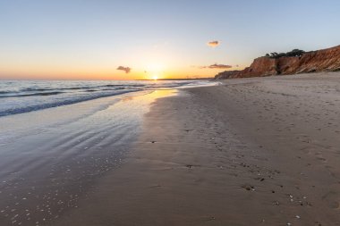 Kızıl kayalıkları ve kayalıkları olan güzel kumlu bir sahil. Günbatımında Praia do Poo Velho, Praia da Falsia, Algarve, Portekiz 'de Atlantik kıyısında sığ dalgalarla birlikte