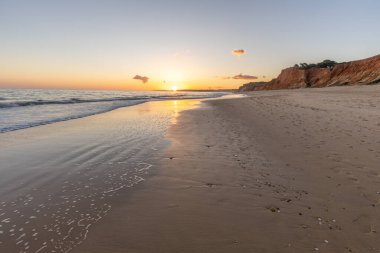 Kızıl kayalıkları ve kayalıkları olan güzel kumlu bir sahil. Günbatımında Praia do Poo Velho, Praia da Falsia, Algarve, Portekiz 'de Atlantik kıyısında sığ dalgalarla birlikte
