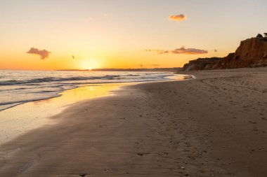 Kızıl kayalıkları ve kayalıkları olan güzel kumlu bir sahil. Günbatımında Praia do Poo Velho, Praia da Falsia, Algarve, Portekiz 'de Atlantik kıyısında sığ dalgalarla birlikte
