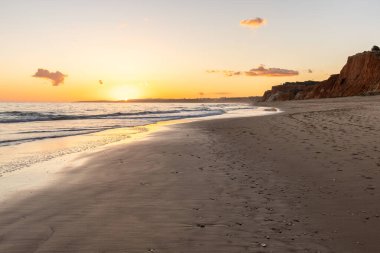 Kızıl kayalıkları ve kayalıkları olan güzel kumlu bir sahil. Günbatımında Praia do Poo Velho, Praia da Falsia, Algarve, Portekiz 'de Atlantik kıyısında sığ dalgalarla birlikte