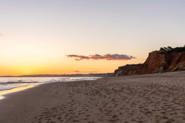 Kızıl kayalıkları ve kayalıkları olan güzel kumlu bir sahil. Günbatımında Praia do Poo Velho, Praia da Falsia, Algarve, Portekiz 'de Atlantik kıyısında sığ dalgalarla birlikte