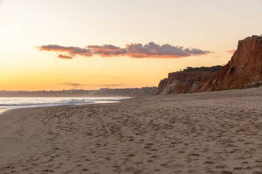 Kızıl kayalıkları ve kayalıkları olan güzel kumlu bir sahil. Günbatımında Praia do Poo Velho, Praia da Falsia, Algarve, Portekiz 'de Atlantik kıyısında sığ dalgalarla birlikte