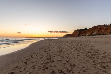Kızıl kayalıkları ve kayalıkları olan güzel kumlu bir sahil. Günbatımında Praia do Poo Velho, Praia da Falsia, Algarve, Portekiz 'de Atlantik kıyısında sığ dalgalarla birlikte