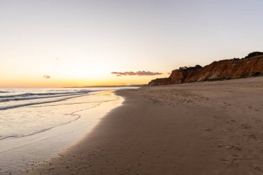 Kızıl kayalıkları ve kayalıkları olan güzel kumlu bir sahil. Günbatımında Praia do Poo Velho, Praia da Falsia, Algarve, Portekiz 'de Atlantik kıyısında sığ dalgalarla birlikte
