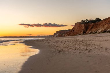 Kızıl kayalıkları ve kayalıkları olan güzel kumlu bir sahil. Günbatımında Praia do Poo Velho, Praia da Falsia, Algarve, Portekiz 'de Atlantik kıyısında sığ dalgalarla birlikte