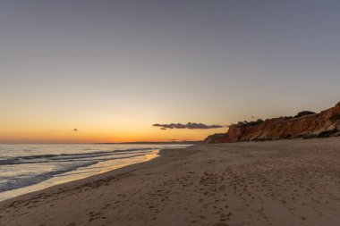 Kızıl kayalıkları ve kayalıkları olan güzel kumlu bir sahil. Günbatımında Praia do Poo Velho, Praia da Falsia, Algarve, Portekiz 'de Atlantik kıyısında sığ dalgalarla birlikte