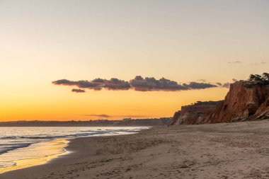 Kızıl kayalıkları ve kayalıkları olan güzel kumlu bir sahil. Günbatımında Praia do Poo Velho, Praia da Falsia, Algarve, Portekiz 'de Atlantik kıyısında sığ dalgalarla birlikte