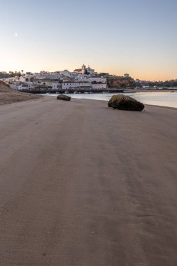 Kumlu bir sahil ve alçak gelgitte limanın gece görüntüsü. Şafakta Ferragudo, Portimo, Algarve, Portekiz 'de aydınlatılmış tarihi bir kasabanın panoramik görüntüsü
