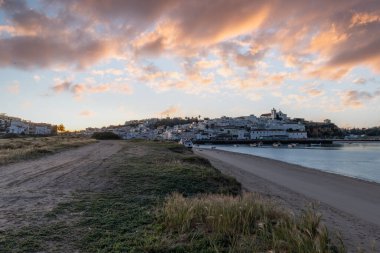 Güneşin doğuşu çayıra, kumlu plaja ve alçak gelgitte limana bakıyor. Sabahın köründe Ferragudo, Portimo, Algarve, Portekiz 'de tarihi bir kasabanın panoramik görüntüsü.