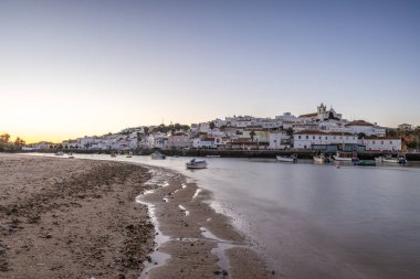 Kumsala bakan bir gün doğumu ve alçak gelgitte liman. Günbatımında Ferragudo, Portimo, Algarve, Portekiz 'de tarihi bir kasabanın panoramik manzarası