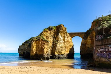 Kumsaldaki sarı kayalar ve uçurumlar. Eski Pinhao Fort Köprüsü 'nün iki uçurumu birleştirdiği deniz kenarında bir koy. Praia da Caldeira plajı, Lagos. Algarve, Portekiz