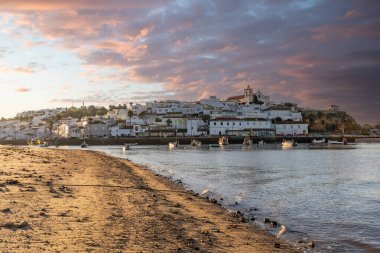 Atlantik Okyanusu 'nda gün doğumu, limanın arkasındaki balıkçı köyünün silueti. Sabah alacakaranlıkta kumsalda. Ferragudo, Portimo, Algarve, Portekiz 'de manzara çekimi