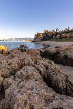 Atlantik 'te gün doğumu, sahilin ortasında bir kale. Limanı koruyan tarihi bir bina. Castelo de Ferragudo, kumlu Praia da Angrinha sahilinde, Portimo, Algarve, Portekiz